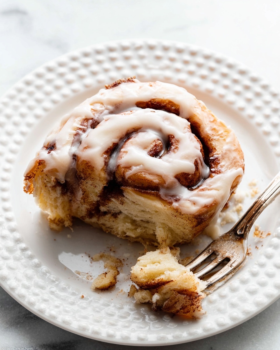 A single slice of cinnamon roll sits on a white plate with a dotted edge design, placed on a white marbled surface. The cinnamon roll has multiple visible layers showing light golden brown dough with swirls of darker cinnamon filling inside. It is topped with a smooth, creamy white glaze that drips softly over the curved layers. Part of the cinnamon roll is eaten, with a few crumbs around. A fork lies on the plate near the bottom right, with traces of cinnamon and glaze on its tines. Photo taken with an iphone --ar 4:5 --v 7