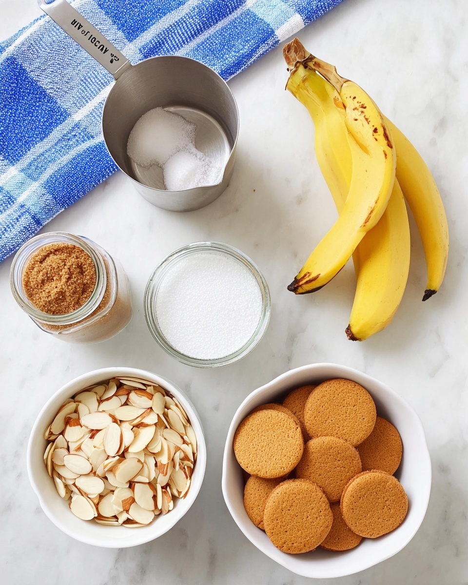 The image shows a white bowl filled with many small round golden-brown cookies on the right side of the frame, with their flat sides and slightly rough textures visible. In the bottom center is another white bowl filled with pale, thinly sliced almonds. To the right of this bowl, two yellow bananas with some dark spots rest on a white marbled surface. Above the almonds is a shiny metal measuring cup filled with white sugar, marked
