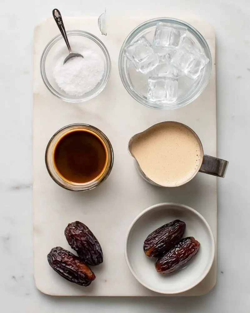 The image shows six ingredients arranged neatly on a white marbled surface. Near the top center, there is a small clear glass bowl filled with coarse white salt, with a metal measuring spoon sticking out. To the right of this bowl, a larger clear glass bowl is filled with clear ice cubes. Below the salt bowl, a short clear glass filled with rich brown espresso with a light foam on top is placed. To the right, a metal measuring cup holds creamy beige unsweetened soy milk, with some small bubbles visible around the edge. Below the espresso glass, two dark brown Medjool dates rest in a small white bowl showing their wrinkled texture. Lastly, at the bottom center of the white marbled surface, a small label identifies vanilla extract, though the extract itself is not shown as a separate container. The overall look is clean and organized. photo taken with an iphone --ar 4:5 --v 7