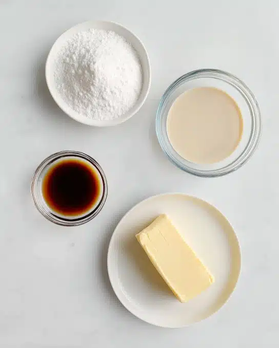 A top view image shows four small white dishes arranged on a white marbled surface. One round dish at the top left is filled with white powdered sugar, fluffy and fine in texture. To its right is a small clear glass bowl holding light beige dairy-free milk, smooth and liquid. Below the powdered sugar is another clear glass bowl containing dark amber vanilla extract, glossy and thin. At the bottom right, a white plate holds a solid, pale-yellow stick of dairy-free butter, smooth with slight surface marks. The layout is neat and minimal. Photo taken with an iphone --ar 4:5 --v 7