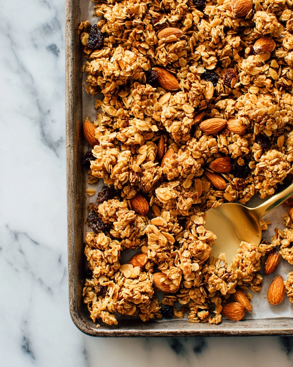 The image shows a baking tray filled with clusters of golden brown granola mixed with whole almonds and some raisins. The granola clusters are uneven in size, with a rough, textured surface of oats and nuts. A golden spoon is partly buried under the granola on the right side of the tray. The tray is placed on a white marbled surface that adds a clean and fresh look to the setting. photo taken with an iphone --ar 4:5 --v 7