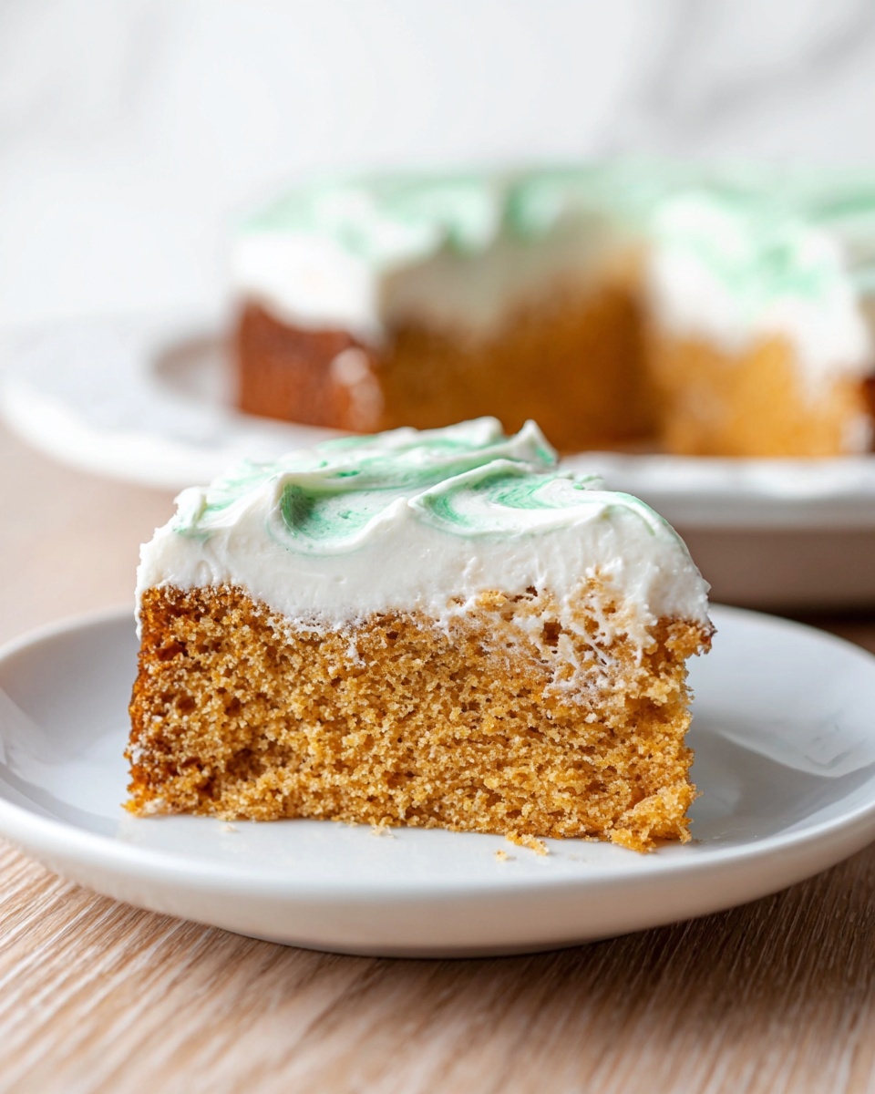 A slice of moist, light-brown cake is shown on a white plate, with two clear layers: the bottom is a soft, spongy cake with a slightly crumbly texture, while the top layer is thick, fluffy white frosting with light green swirls smoothly spread across the surface. The slice is sitting on a wooden surface with a white marbled background. In the back, the rest of the cake in a white dish is slightly out of focus. photo taken with an iphone --ar 4:5 --v 7