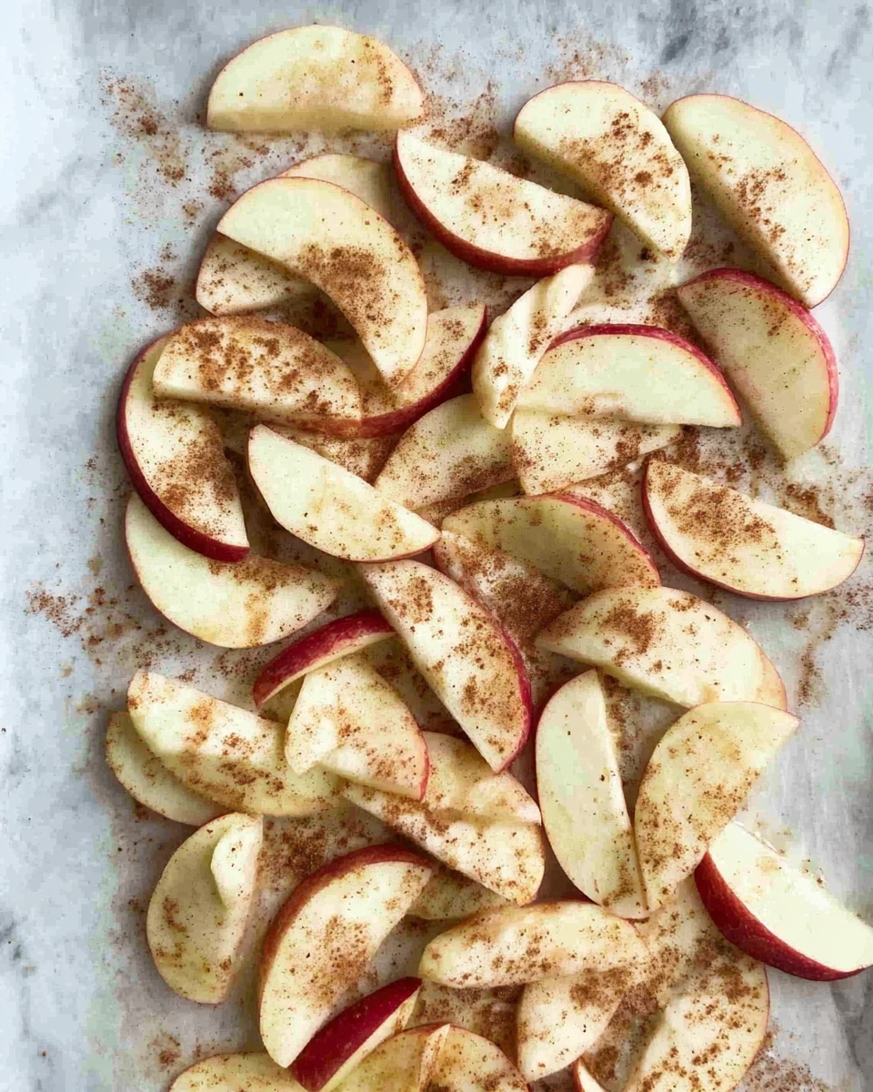 The image shows a single layer of apple slices spread out evenly on a white marbled baking sheet or tray. The apple slices are thin, pale yellow with red skin edges, and lightly sprinkled with a brown powder that looks like cinnamon. The white marbled background adds brightness to the scene. photo taken with an iphone --ar 4:5 --v 7