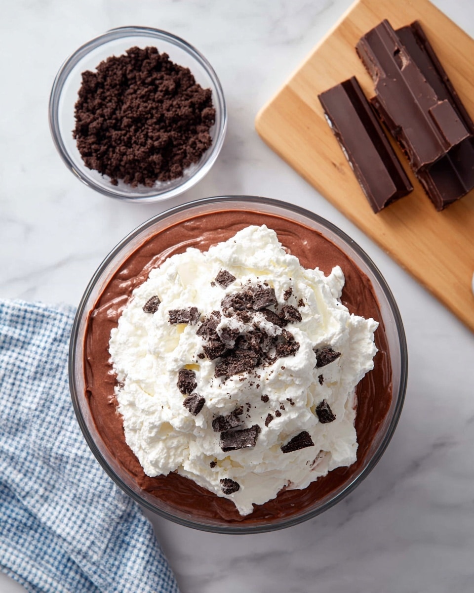 A clear glass bowl shows three layers: the bottom layer is smooth, thick chocolate pudding with a dark brown color. On top of it, there are small chunks of crushed dark chocolate cookies scattered around the edges. The top layer is a big mound of fluffy white whipped cream that covers most of the cookie bits. Nearby, a small clear bowl holds extra dark chocolate cookie crumbs, and three milk chocolate bars rest on a light wooden cutting board. The whole scene is set on a white marbled surface with a corner of a blue and white checkered cloth visible. Photo taken with an iphone --ar 4:5 --v 7