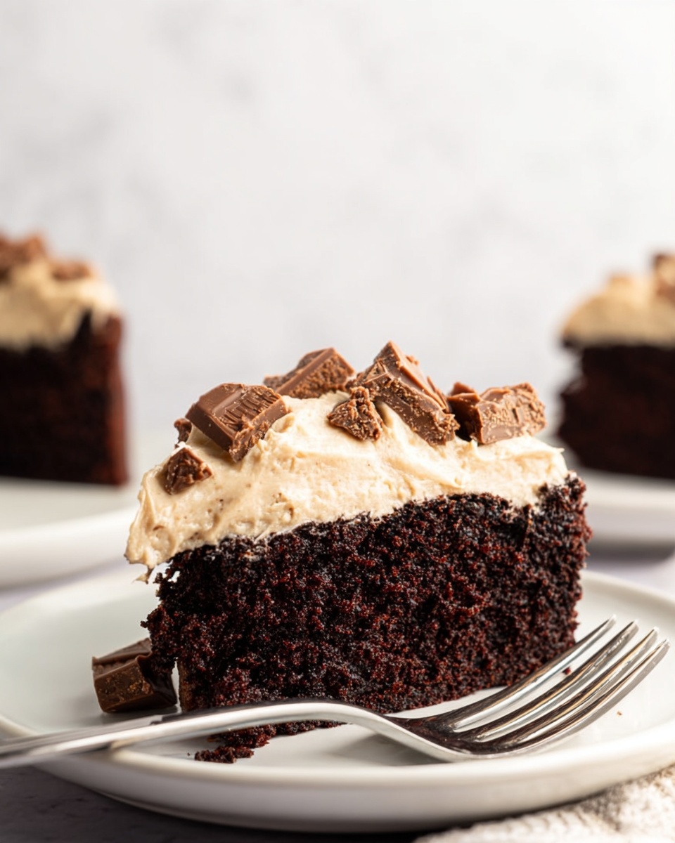 The image shows a thick slice of dark brown chocolate cake with a moist texture, topped with a thick layer of light beige frosting that looks creamy and smooth. Small pieces of chopped chocolate candy bar are scattered on top of the frosting, adding texture and color contrast with their light brown and chocolate shades. The slice is placed on a simple white plate with a shiny silver fork beside it. The background is a soft white marbled surface that highlights the rich colors of the cake, with another blurred slice of cake in the background. The photo was taken with an iphone --ar 4:5 --v 7