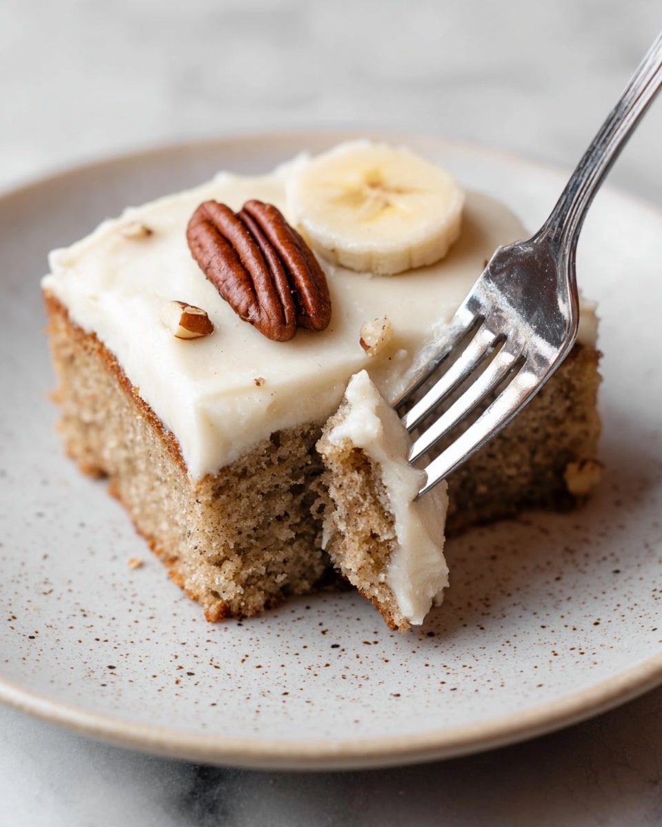 A square piece of light brown banana cake with a soft texture sits on a white plate with small speckles on it. The cake has a thick, smooth layer of creamy off-white frosting on top, decorated with a pecan nut placed near the center and a round, slightly glossy slice of banana resting beside it. A shiny silver fork is inserted into the side of the cake, lifting a small piece covered in frosting. The surface under the plate is a white marbled texture. Photo taken with an iphone --ar 4:5 --v 7
