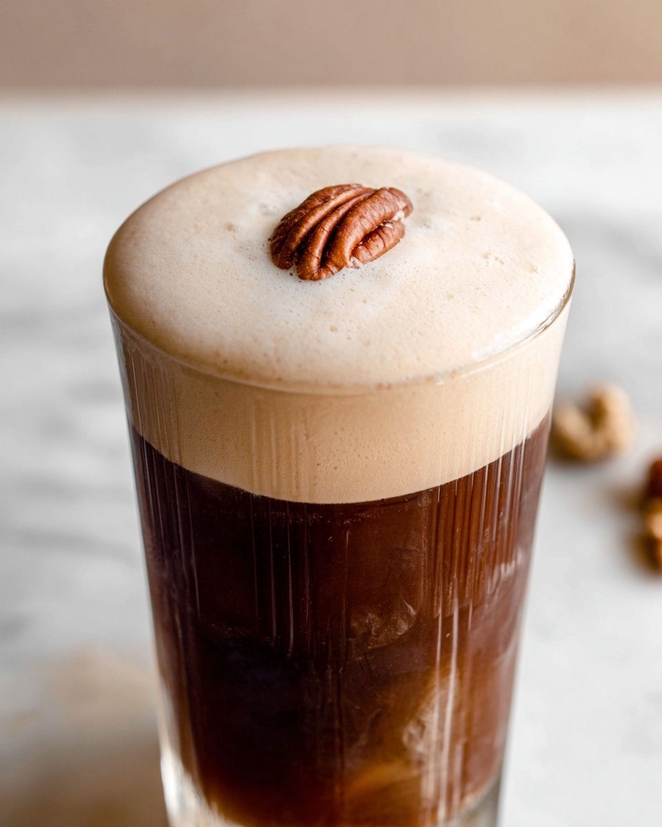 A tall transparent glass filled with three visible layers: the bottom layer is light brown with a smooth texture, the middle layer is dark brown coffee with streaks running vertically down the glass, and the top layer is a thick, frothy, creamy foam unevenly spread over the surface. The glass is placed on a white round coaster, set on a white marbled texture surface. The background is softly blurred with a jar of syrup and some nuts partially visible. Photo taken with an iphone --ar 4:5 --v 7