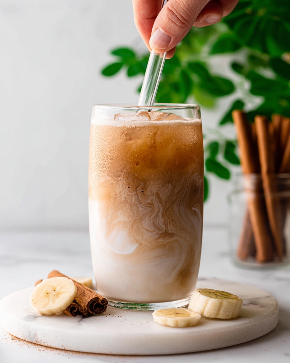 A clear glass filled with a swirled drink has three visible layers: a light brown layer at the bottom with ice, a medium brown middle layer mixing with a creamy white top layer, creating a smooth blend. A woman's hand stirs the drink with a glass straw from above. The glass sits on a round white marble surface with two banana slices next to it. In the background, there are cinnamon sticks in a clear container and green leaves adding a fresh touch. The overall setting is bright with a white marbled texture photo taken with an iphone --ar 4:5 --v 7