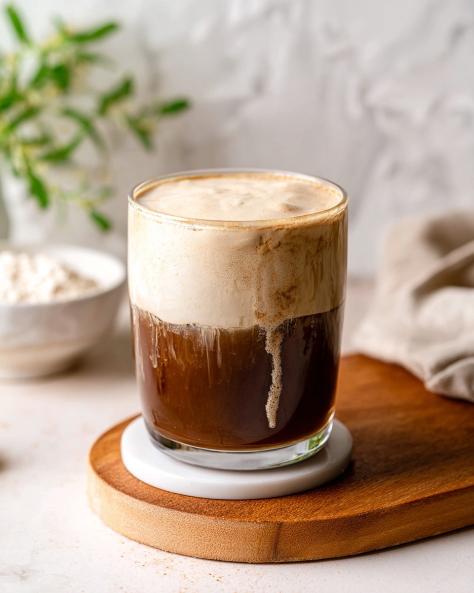 A clear glass sits on a white round coaster on a wooden board, filled with a dark brown liquid on the bottom and a thick, creamy beige foam layer on top, about one third of the glass, with the foam slightly dripping down the sides. The background is a soft, white marbled texture with small green leaves and a small white bowl filled with powder blurred behind. The light softly brightens the drink’s smooth textures. Photo taken with an iphone --ar 4:5 --v 7