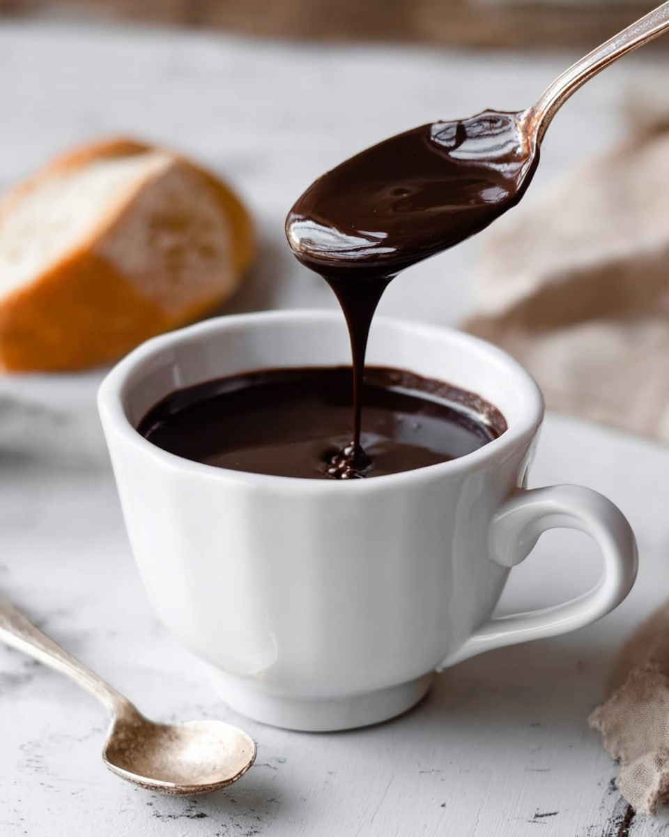 A white cup on a white marbled surface holds thick dark brown chocolate sauce, almost filling the cup. Above the cup, a spoon with a long handle drips the shiny chocolate sauce back into the cup, showing a smooth and glossy texture. Next to the cup, there is a small silver spoon lying on the surface. In the background, there is a blurred piece of bread resting on the white marbled surface. photo taken with an iphone --ar 4:5 --v 7
