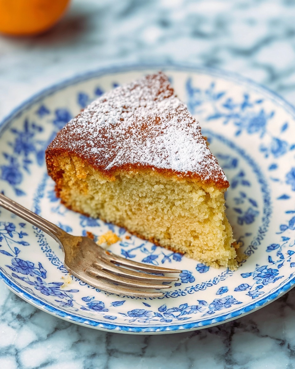 A single slice of cake with one visible layer sits on a white plate decorated with blue floral patterns. The cake layer is light golden brown with a soft, slightly crumbly texture. The top of the cake has a slightly darker brown color with a light dusting of white powdered sugar scattered unevenly. A silver fork rests diagonally on the plate near the bottom left of the cake slice. The plate is placed on a surface with a white marbled texture. Photo taken with an iphone --ar 4:5 --v 7