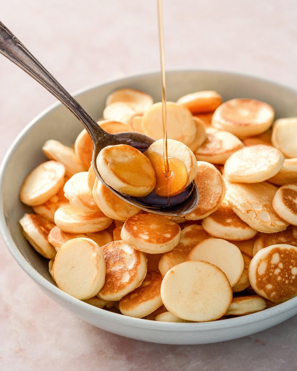 A bowl filled with many small, round, light beige mini pancakes stacked unevenly, showing their soft and slightly spongy texture. A silver spoon rests inside the bowl, holding several mini pancakes, while golden syrup or honey is being poured over them, creating a shiny and glossy layer that pools slightly in the spoon. The bowl is white, placed on a white marbled surface with soft light that highlights the warm tones of the pancakes and syrup. photo taken with an iphone --ar 4:5 --v 7