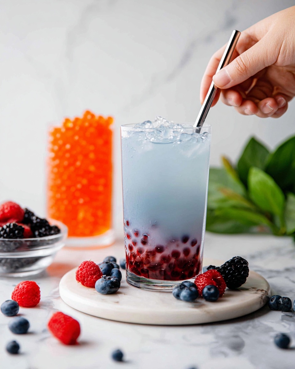 A clear glass filled with a light blue drink layered with small dark red tapioca pearls at the bottom, topped with ice cubes, is being stirred by a woman's hand holding a metal straw from the right side. The glass sits on a round white marble plate with scattered fresh berries around it, including blueberries, raspberries, and blackberries. In the background, there is a tall clear glass filled with bright orange spheres and a green leafy plant, all set against a white marbled surface. photo taken with an iphone --ar 4:5 --v 7