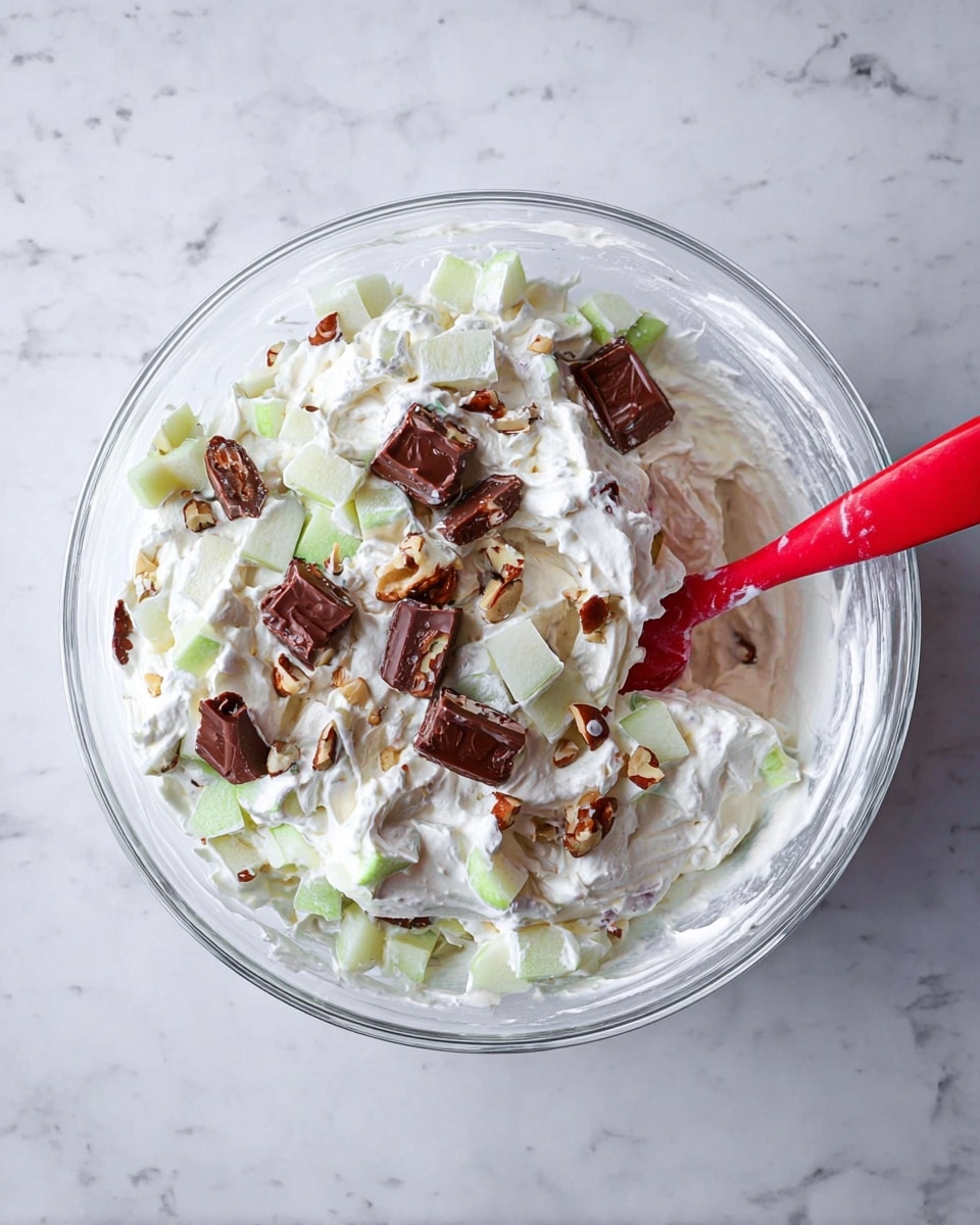 A clear glass bowl sits on a white marbled surface filled with a creamy mixture made of white whipped cream and small light green apple pieces, along with some pale yellow chunks, all mixed evenly. On top of the creamy base, there are several small pieces of chocolate candy with visible nut filling, adding a dark brown and light tan contrast. A red spatula is partially dipped into the mixture on the right side of the bowl. photo taken with an iphone --ar 4:5 --v 7