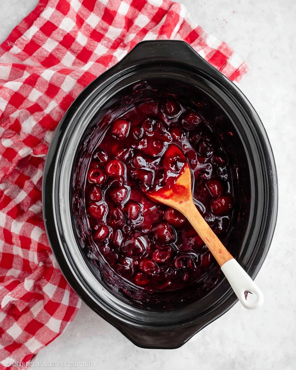 The image shows a black slow cooker filled with a thick, glossy cherry sauce that is dark red with visible whole cherries and chunks, giving it a textured look. Inside the cooker, a wooden spoon with a white tip is resting in the sauce, coated with some of the cherry mixture. To the left of the cooker, there is a red and white checkered cloth lying on a white marbled surface that covers the whole background. photo taken with an iphone --ar 4:5 --v 7