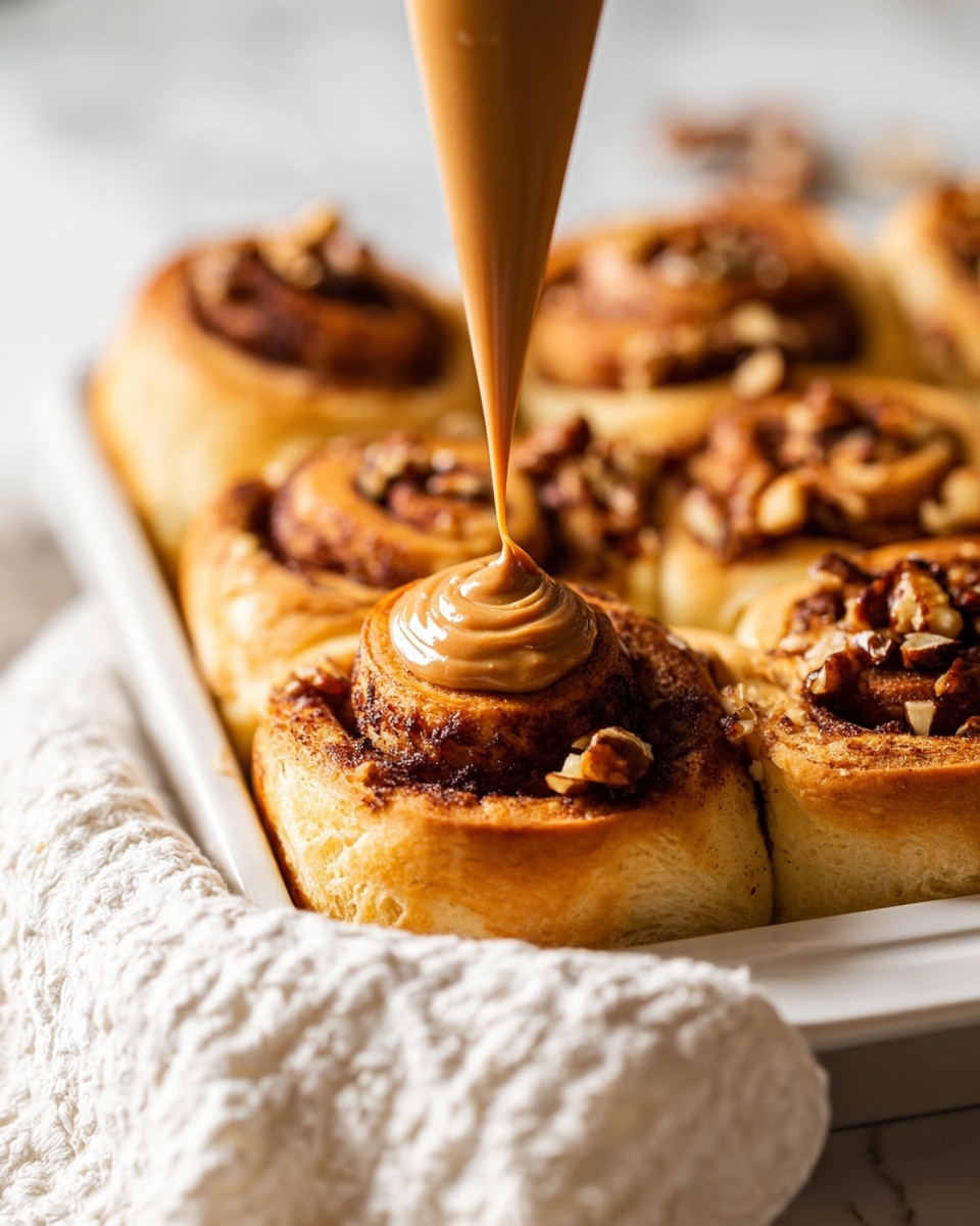 A close-up image shows a tray of golden-brown cinnamon rolls with visible nuts on top, arranged closely together in a baking dish with a white marbled surface beneath. One cinnamon roll is centered as a thick, light brown caramel-like sauce is being squeezed in a smooth swirl from a piping bag onto its top. The rolls have a soft, fluffy texture with darker brown cinnamon swirls inside, and the edges are slightly crisp. A white cloth with a textured pattern is placed in the foreground, adding to the cozy atmosphere. Photo taken with an iphone --ar 4:5 --v 7