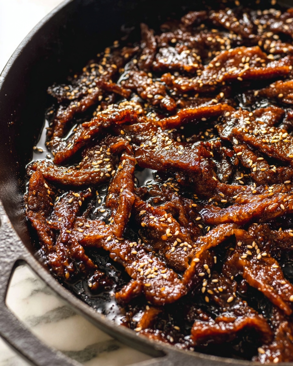 The image shows many thin strips of dark brown meat cooked in a black cast iron pan. The meat has a shiny, sticky texture from a thick sauce and is sprinkled evenly with small white sesame seeds. The pan handle is visible at the bottom left, and the pan sits on a white marbled surface. The focus is close up, showing the rich texture of the meat and sesame seeds clearly. photo taken with an iphone --ar 4:5 --v 7