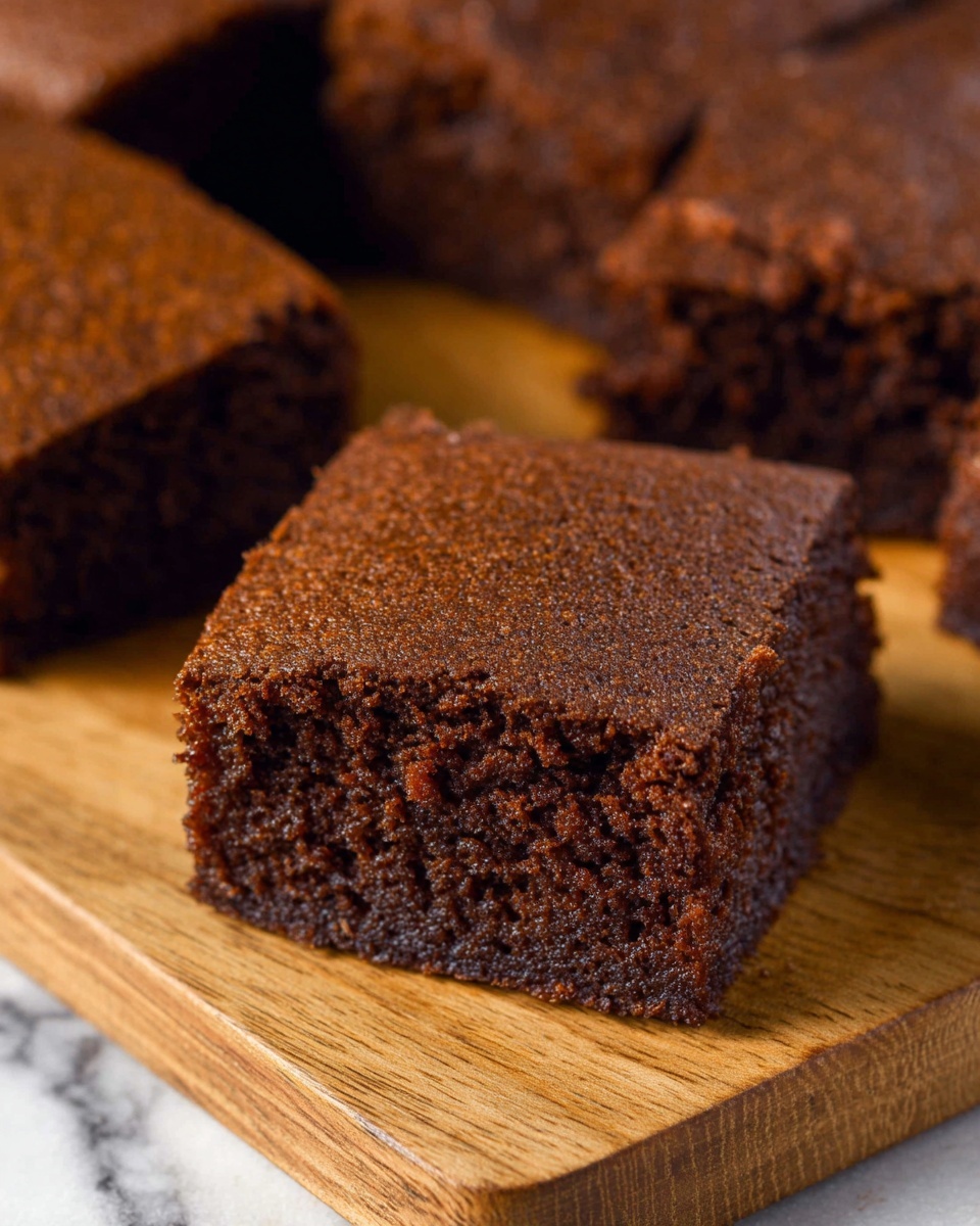 The image shows a close-up of a single square piece of dark brown cake with a textured top and sides, appearing moist and soft. It sits on a light brown wooden board with several more square pieces of the same cake arranged in the background. The scene is set on a white marbled surface, and the cake pieces have a slightly rough, homemade look. Photo taken with an iphone --ar 4:5 --v 7