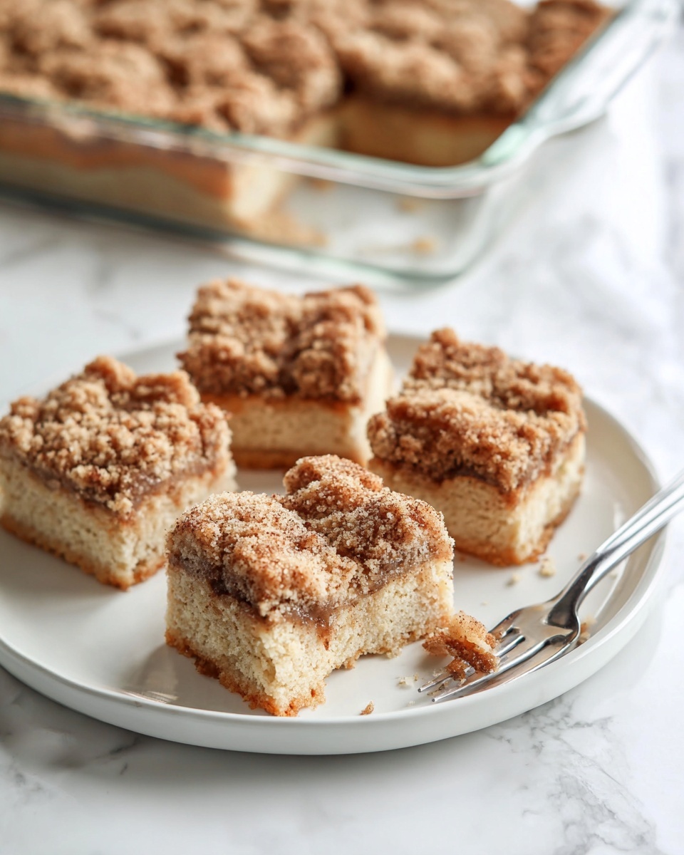 The image shows five square pieces of crumb cake on a white plate, each with two layers: a light beige soft cake base and a thick crumb topping layer in a golden brown color with a crunchy texture. Pieces have a rough, crumbly top with specks of darker cinnamon color scattered throughout. One piece is partially eaten with a small bite taken out of one corner, placed beside a silver fork on the plate. The scene is set on a white marbled surface with a glass baking dish of more crumb cake blurred in the background. photo taken with an iphone --ar 4:5 --v 7