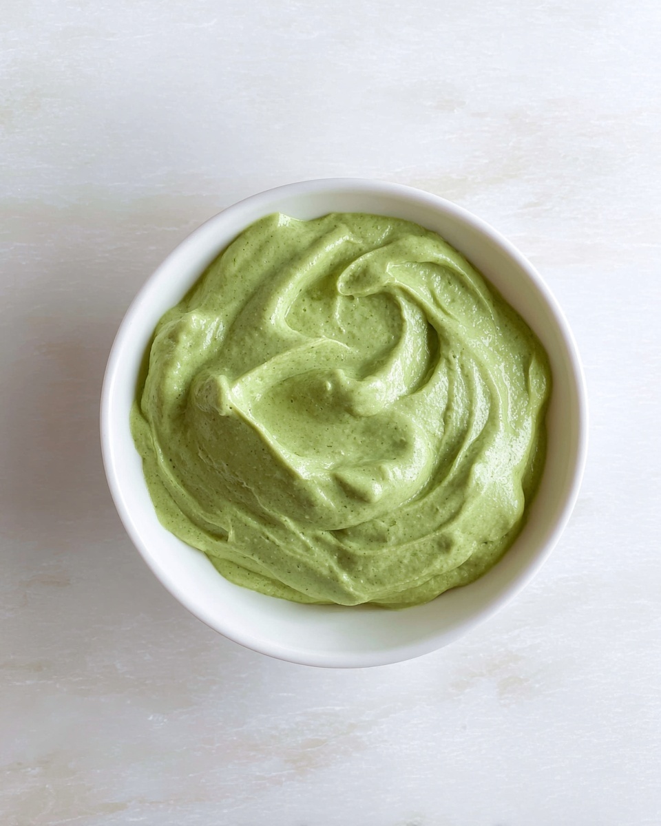 A white bowl filled with a thick, creamy green sauce with a smooth and slightly fluffy texture. The sauce has gentle swirls and soft peaks, showing a fresh and rich consistency. The bowl sits on a white marbled surface. photo taken with an iphone --ar 4:5 --v 7