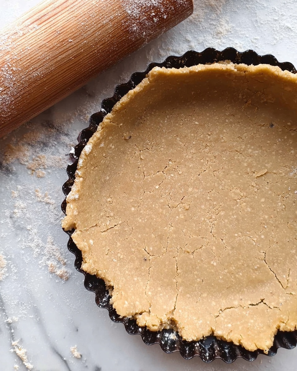 A close-up top view of a pie crust inside a black fluted tart pan. The crust dough is light brown with a rough texture and some cracks, extending unevenly over the edges of the pan. There are small specks of flour dusted on the surface of the dough. A wooden rolling pin with some flour on it lies diagonally on a white marbled textured surface beneath the tart pan. photo taken with an iphone --ar 4:5 --v 7