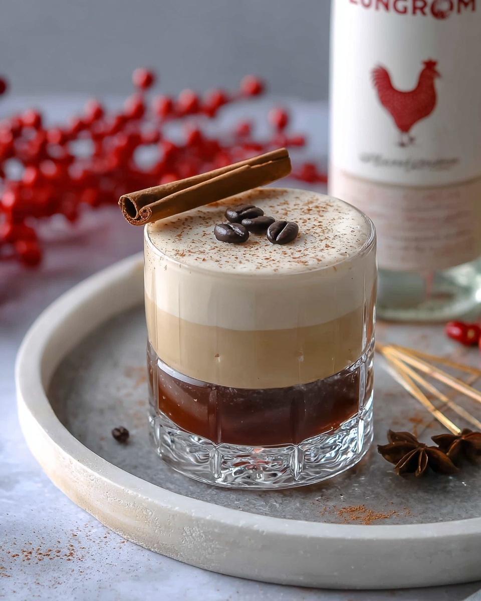 A clear glass holds a two-layer drink with a deep brown layer on the bottom and a thick creamy light beige layer on top. Three dark coffee beans sit in the center of the top layer, lightly dusted with fine specks of spice. A cinnamon stick rests horizontally on the rim of the glass. The glass is placed on a round white tray with a subtle texture, along with star anise and thin caramel-like elements. In the background, a white bottle with red text and a chicken illustration stands beside some blurred red beads on the white marbled surface. photo taken with an iphone --ar 4:5 --v 7