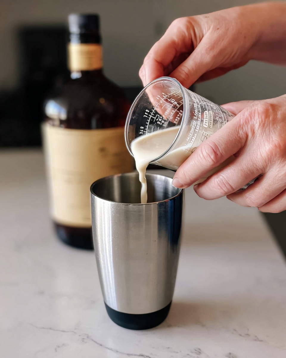 The image shows a close-up of a person's two hands pouring a light cream liquid from a small clear measuring cup into a tall silver metal cup with a black rim, held steady on a white marbled surface. Behind the hands, there is a dark brown glass bottle with a gold and black cap, slightly blurred in the background. The scene is focused on the liquid stream flowing from the measuring cup into the metal cup, creating a simple and clean visual with soft natural light. photo taken with an iphone --ar 4:5 --v 7