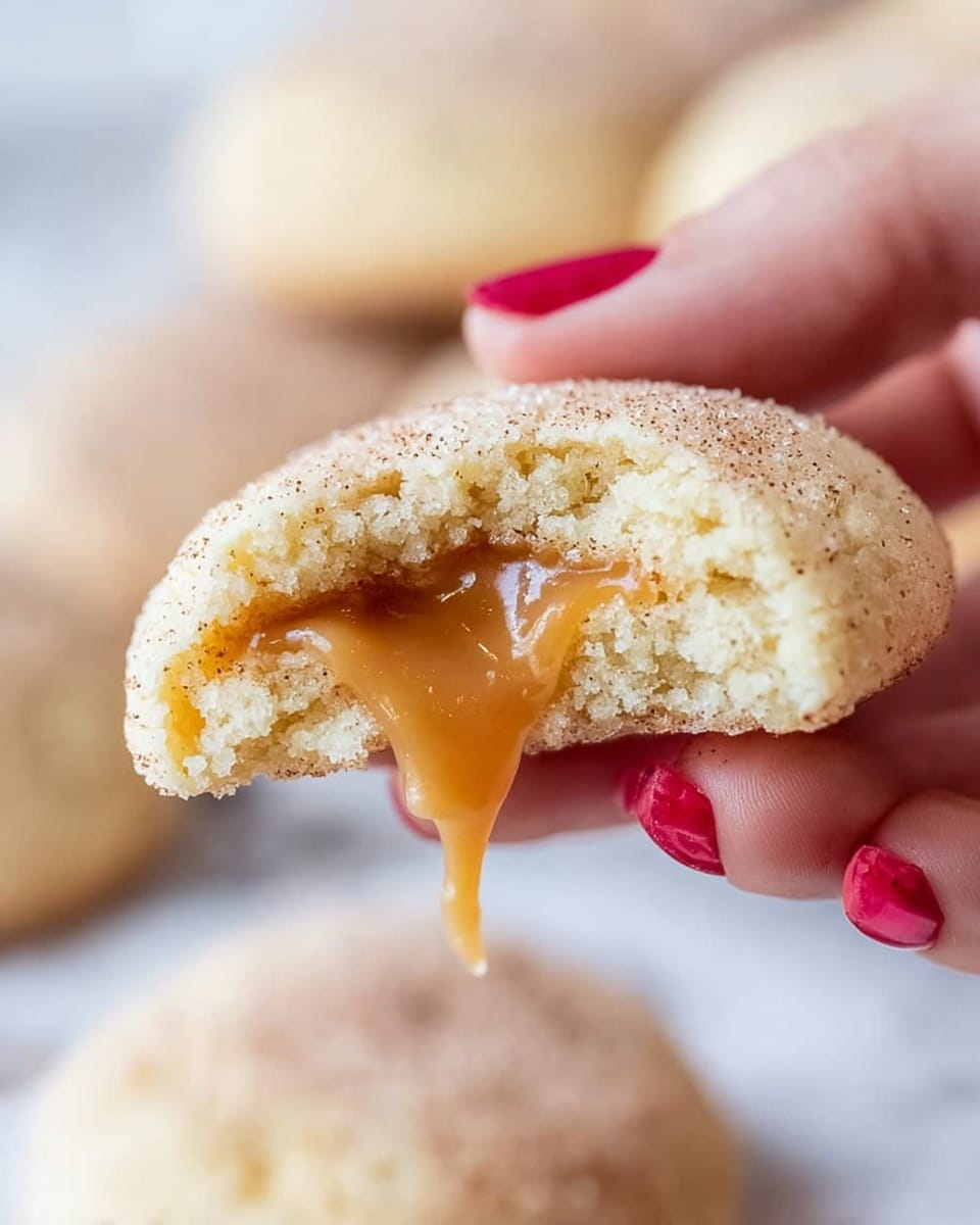 A close-up view shows a soft, round cookie being gently held by a woman's hand with red polished nails. The cookie has a light golden-brown outer layer with a slightly grainy texture and is broken open in the middle, revealing a gooey, smooth caramel filling that stretches between the two halves. The soft inside of the cookie is pale and fluffy. The background is a white marbled texture with more cookies softly blurred in the distance. photo taken with an iphone --ar 4:5 --v 7