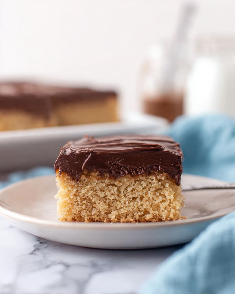 A square piece of cake with two layers sits on a white plate against a white marbled surface. The bottom layer is light brown, soft, and crumbly. The top layer is dark brown chocolate frosting, smooth and thick. In the blurred background, another piece of cake with chocolate frosting is visible, alongside two small glass jars and a soft blue cloth on the right side. Photo taken with an iphone --ar 4:5 --v 7