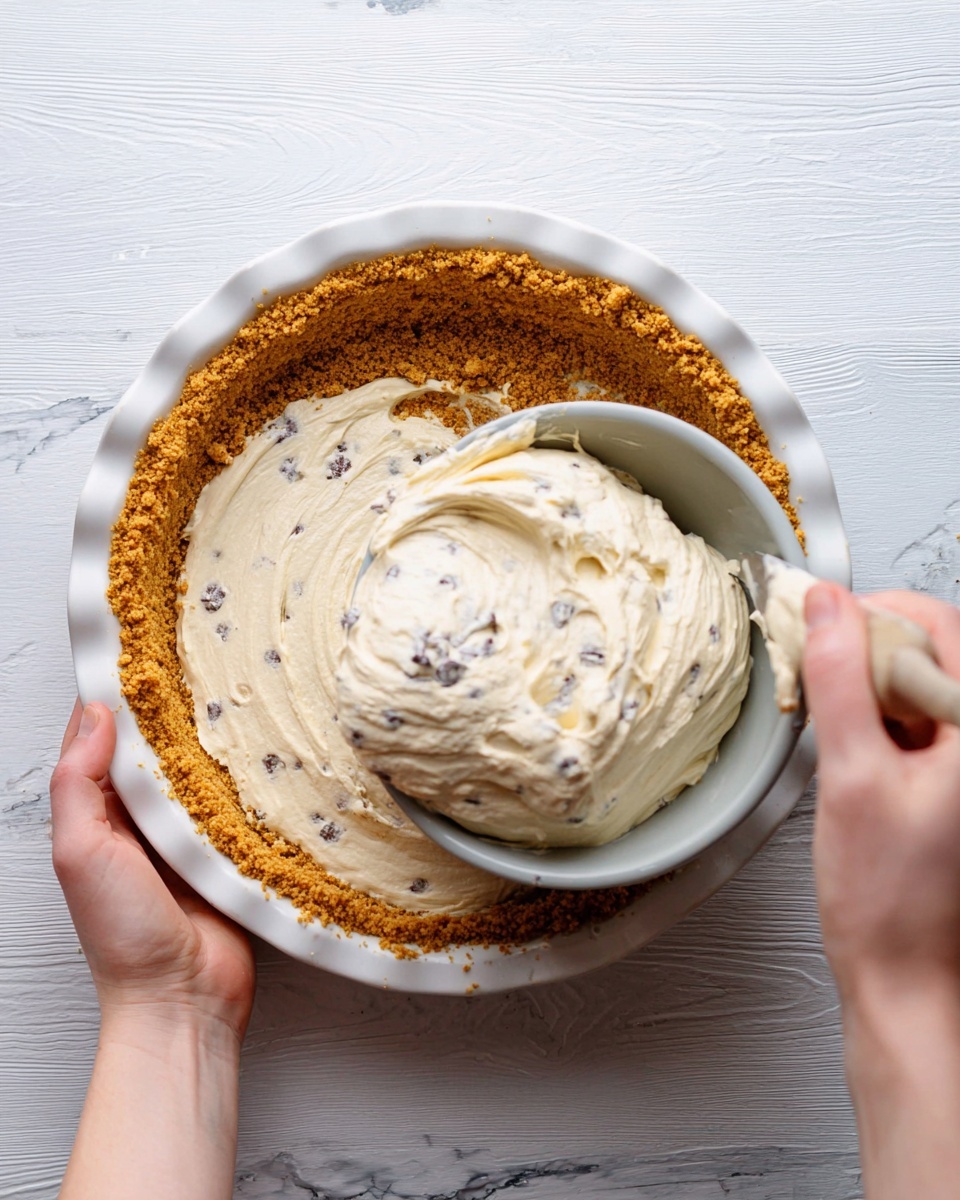 A white pie dish holds a golden brown, crumbly crust pressed evenly with a slightly rough texture, forming the bottom and sides. A woman's hand is seen spreading a thick, creamy, off-white filling with small dark chocolate chips inside, scooped from a light grey bowl above. The filling has a smooth and soft texture with a few folds and peaks as it's being transferred into the crust. The scene is set on a white marbled surface, adding a clean and bright background to the process. photo taken with an iphone --ar 4:5 --v 7