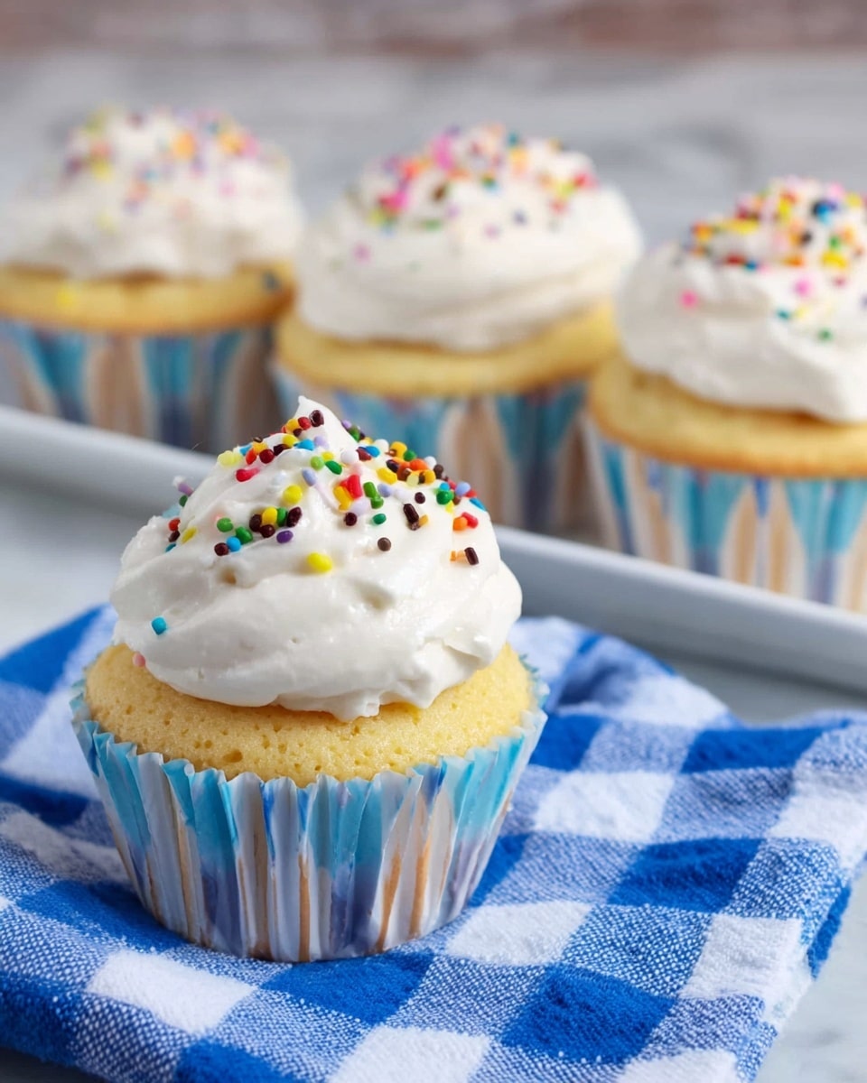 The image shows a close-up of a single cupcake in the front with three more cupcakes blurred in the background, all placed on a white tray on a white marbled surface. Each cupcake has one layer of light yellow cake with a slightly rough texture, wrapped in blue and white striped paper liners. On top of each cupcake is a smooth, thick layer of white whipped frosting, which is decorated with small, colorful sprinkles in red, brown, yellow, green, blue, and pink. The cupcake in front rests on a blue and white checkered cloth, adding a cozy touch. Photo taken with an iphone --ar 4:5 --v 7