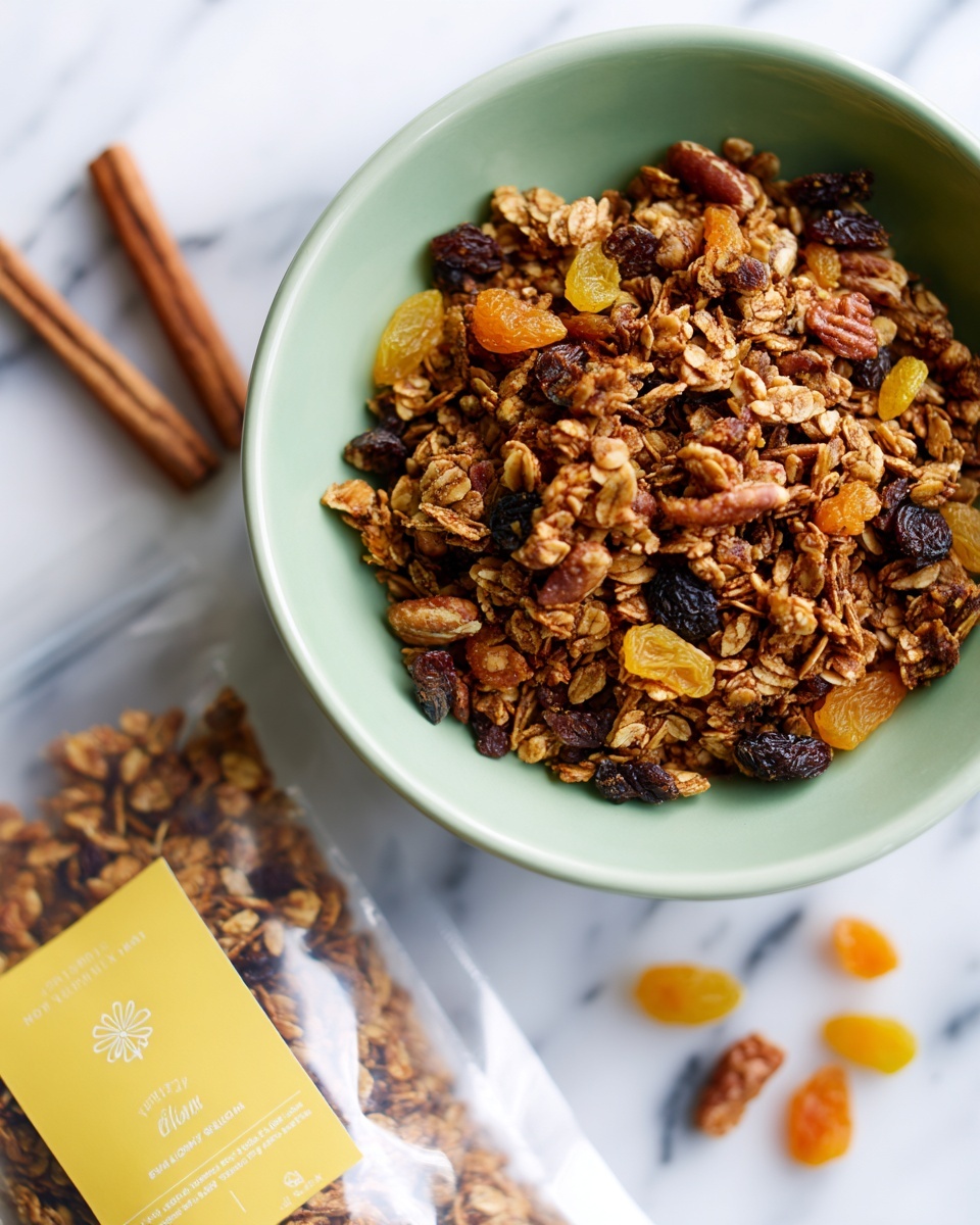 A close-up image of a light green bowl filled with granola made of oats, nuts, and dried fruits like raisins and small apricot chunks. The bowl is placed on a white marbled surface. Some granola pieces are scattered around the bowl beside a clear plastic package with a yellow label featuring cinnamon sticks on it. The granola shows a mix of tan and brown colors with crunchy textures. Photo taken with an iphone --ar 4:5 --v 7