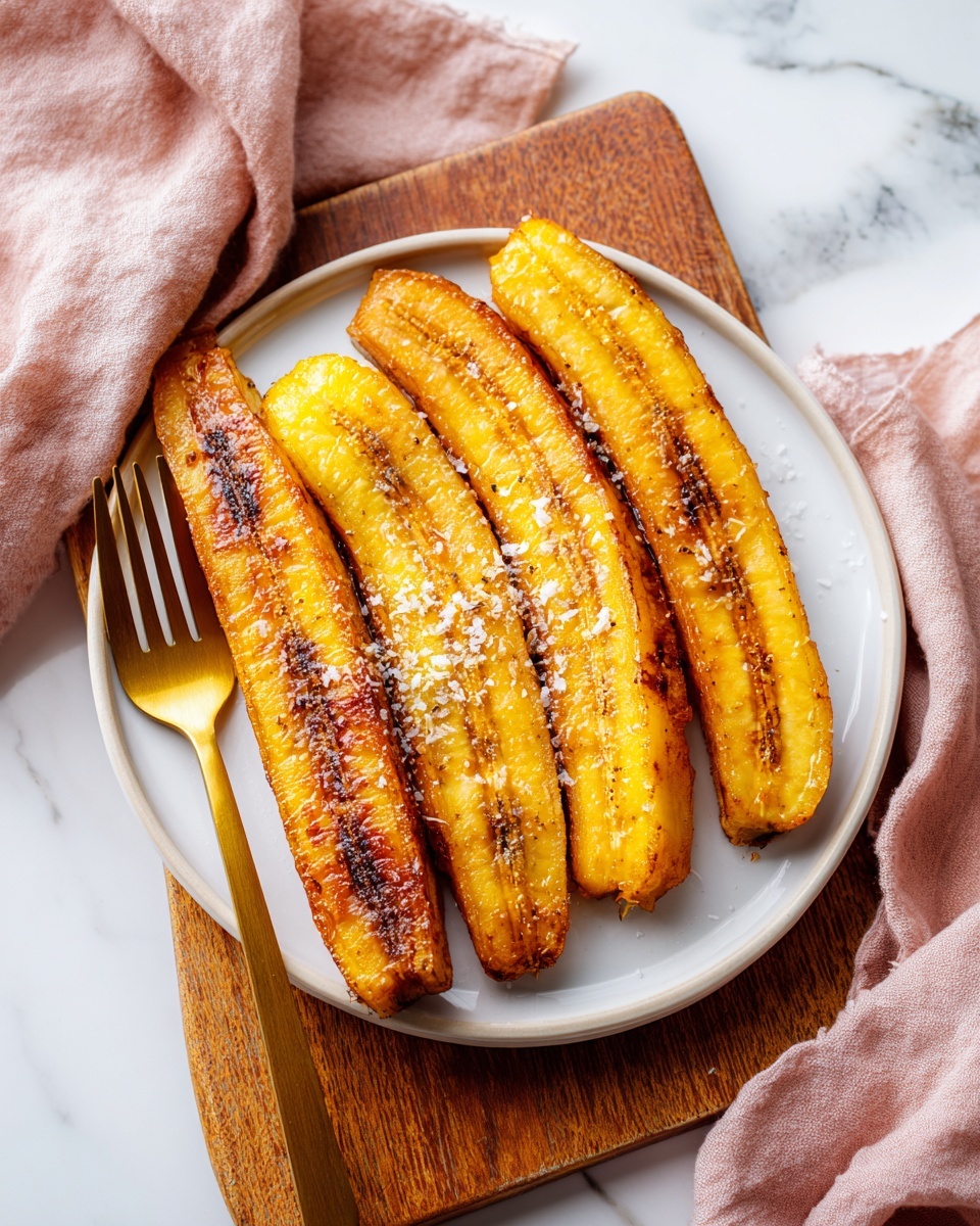 The image shows a white plate with four long, cooked plantains arranged side by side in a fan shape. Each plantain has a golden-brown, slightly crispy surface with darker caramelized edges and a light sprinkle of coarse salt. The plate is placed on a light wooden cutting board with a natural edge, resting on a white marbled surface. Next to the plate is a bronze fork lying diagonally on the cutting board, with a crumpled soft pink cloth draped behind it, adding texture and color to the scene. Photo taken with an iphone --ar 4:5 --v 7