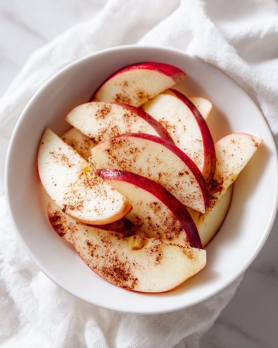 Several thin apple slices sprinkled with cinnamon are spread in a white bowl lined with a white cloth, showing the apple skins dark red around the edges and the apple flesh a creamy beige with small brown cinnamon spots. The bowl sits on a white marbled surface. Photo taken with an iphone --ar 4:5 --v 7
