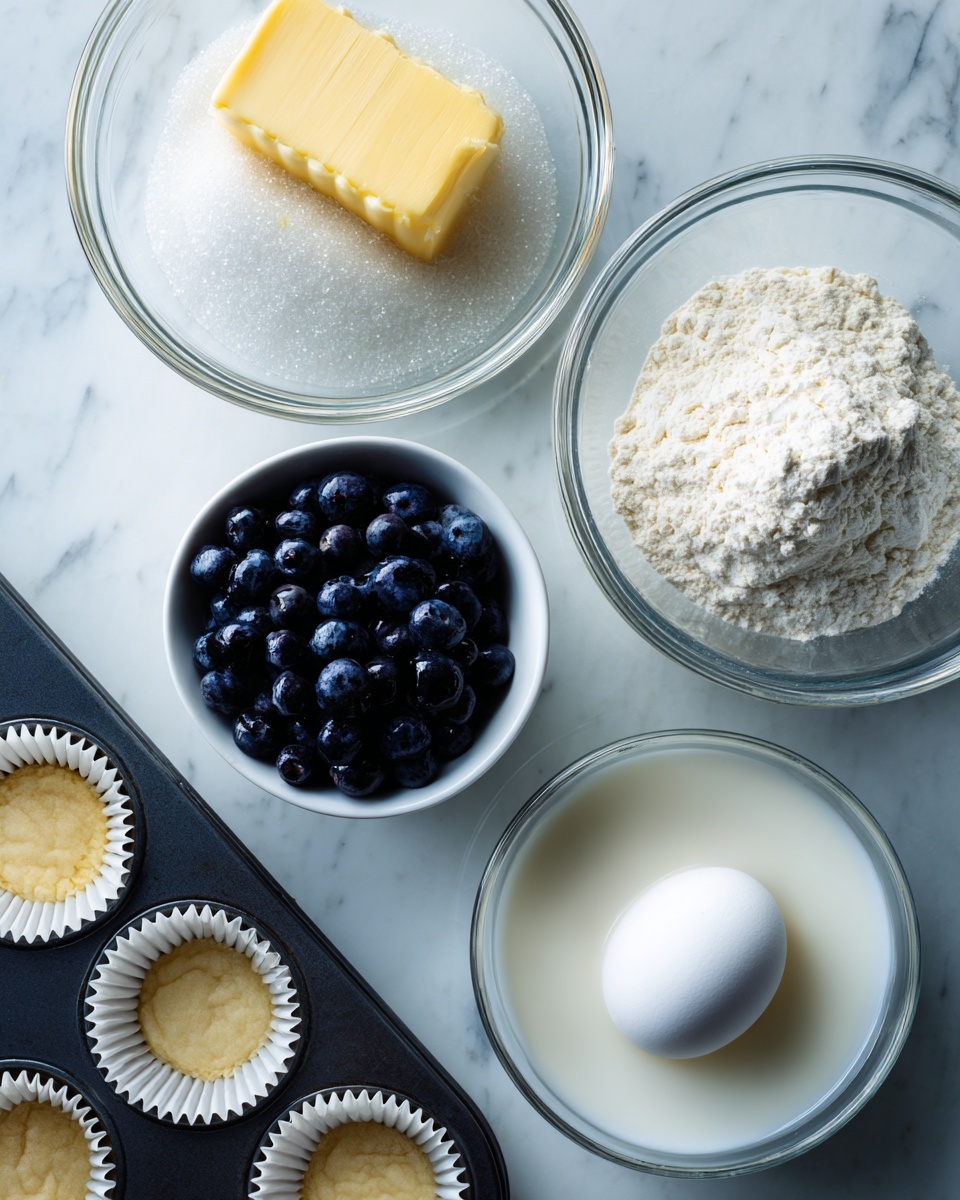 A glass bowl at the top left contains a yellow stick of butter partially covered by white sugar grains, showing a grainy and creamy texture together. Below it, another glass bowl holds white flour with a slightly powdery look. To the top right, a small white bowl is filled tightly with dark, shiny blueberries giving a smooth round texture. Next to it, there is a white bowl with some milk that looks smooth and shiny. At the bottom left, a dark muffin tray holds white paper liners, with a white egg sitting in one of the liners, showing a smooth, clean surface. All items are placed on a white marbled surface. Photo taken with an iphone --ar 4:5 --v 7