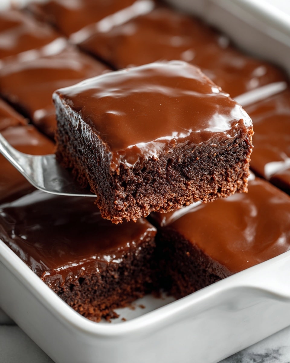 A close-up of a square piece of chocolate cake with two layers resting on a spatula over a clear glass baking dish. The bottom layer is dark, moist, and dense cake with a rough texture. The top layer is a smooth, light brown chocolate frosting that covers the entire surface. The cake is cut into neat squares, with one piece removed to show the empty space in the baking dish. The background shows a soft white marbled texture. Photo taken with an iphone --ar 4:5 --v 7