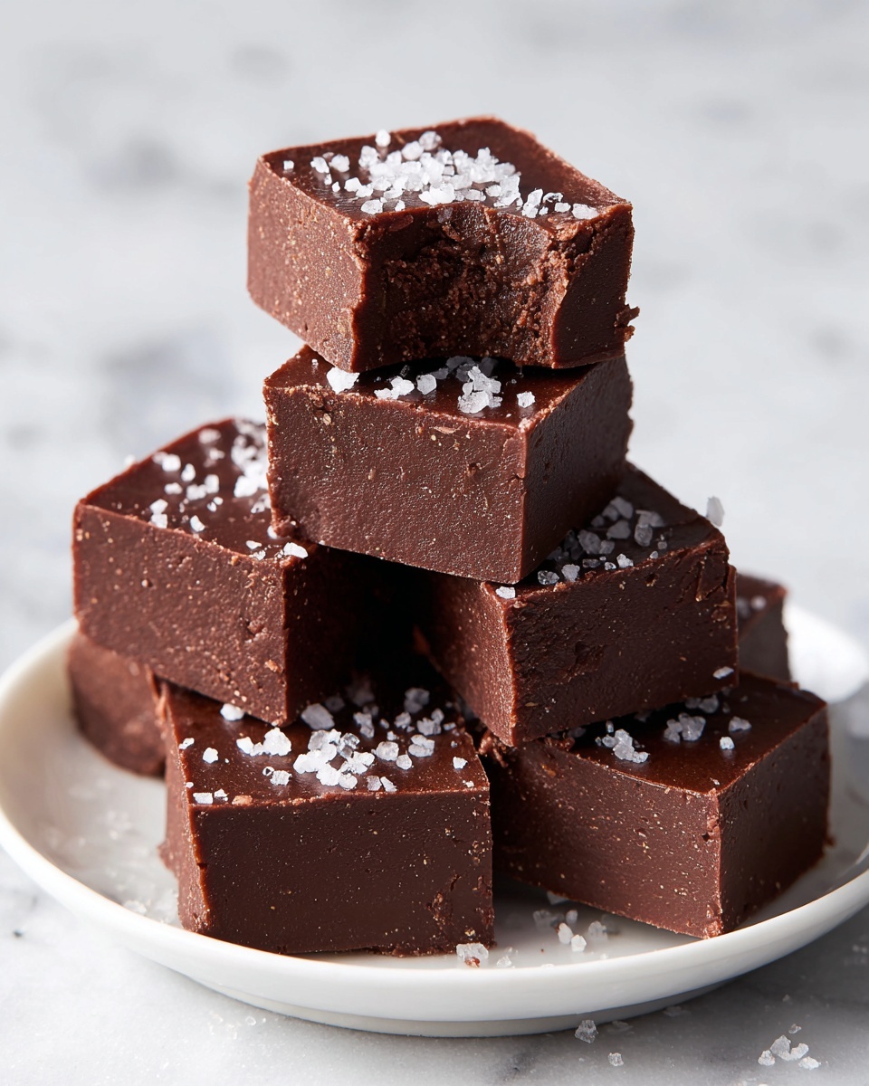 A stack of small, square chocolate fudge pieces is placed on a white plate, each piece topped with white sea salt flakes. The fudge has a smooth, slightly shiny dark brown top layer and a crumbly textured bottom layer visible where some pieces have small bites taken out. The white marbled surface under the plate adds a clean and bright contrast to the rich brown tones of the fudge. photo taken with an iphone --ar 4:5 --v 7