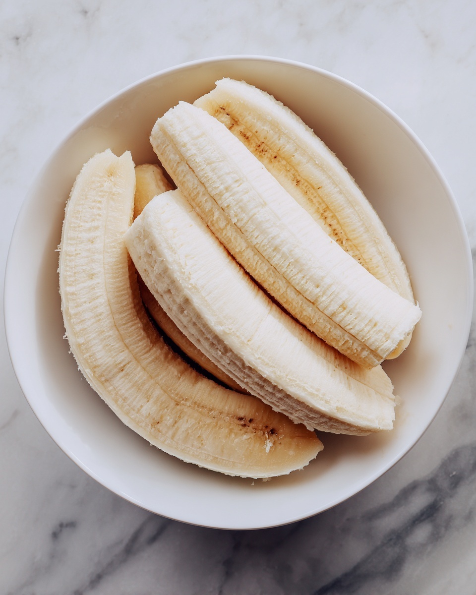 The image shows a white bowl with several peeled bananas inside. The bananas are whole, placed on top of each other in a loose pile, and have a soft, pale yellow color with some light brown spots. The texture looks slightly fibrous and moist, showing the natural look of peeled bananas. The background is a white marbled surface. photo taken with an iphone --ar 4:5 --v 7